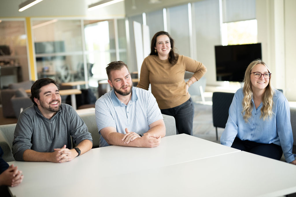 Two BerkleyNet employees standing in a bright office space. Four BerkleyNet employees sitting in a bright office space, a welcoming and professional team dedicated to fast and easy workers compensation insurance solutions.